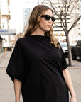 A close up of a woman wearing a black boatneck dress with asymmetrical sleeves and hem by Malaika New York