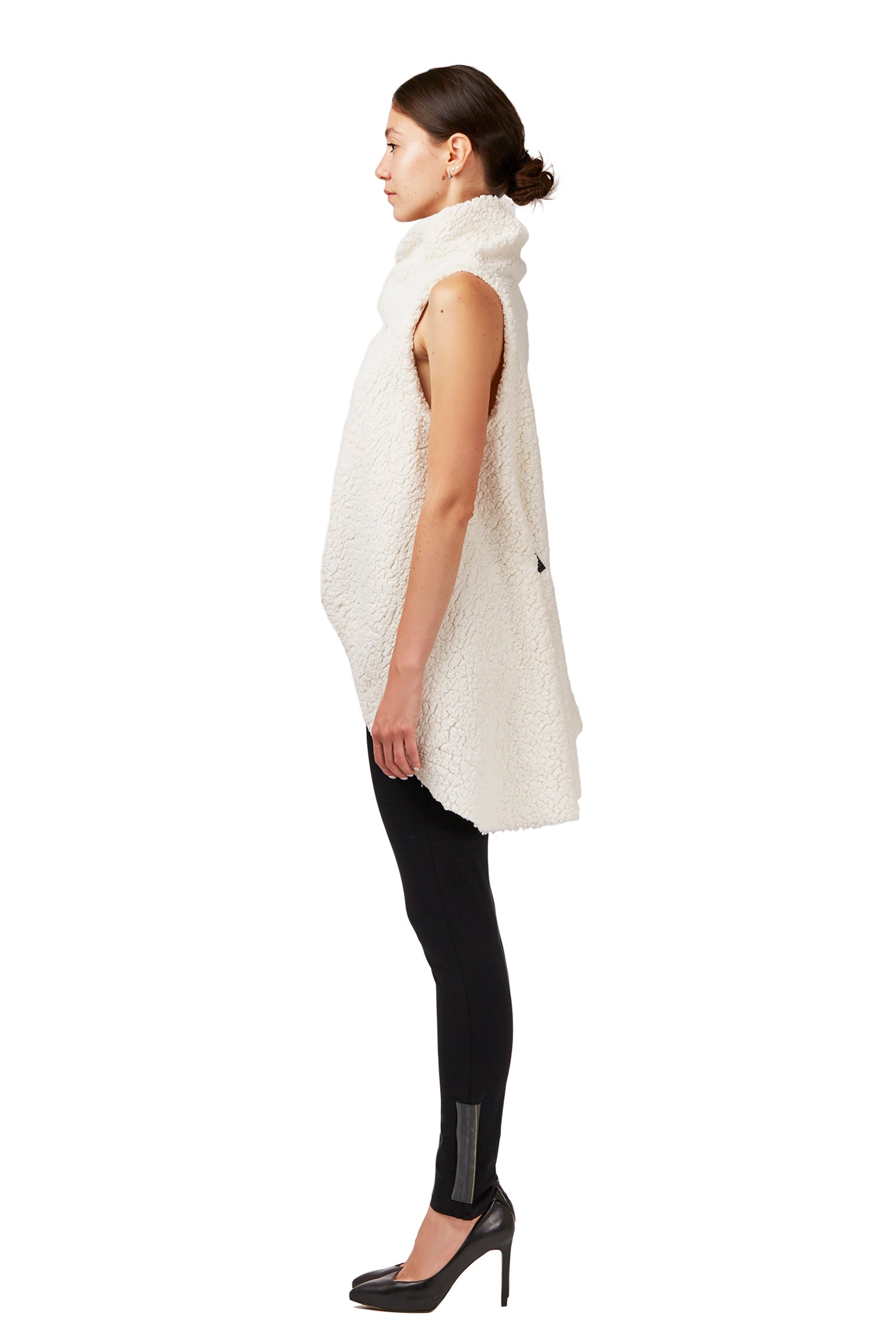 A woman standing with her side turned  wearing a white faux fur asymmetrical vest with black snaps &amp; a pair of black organic cotton leggings by Malaika New York