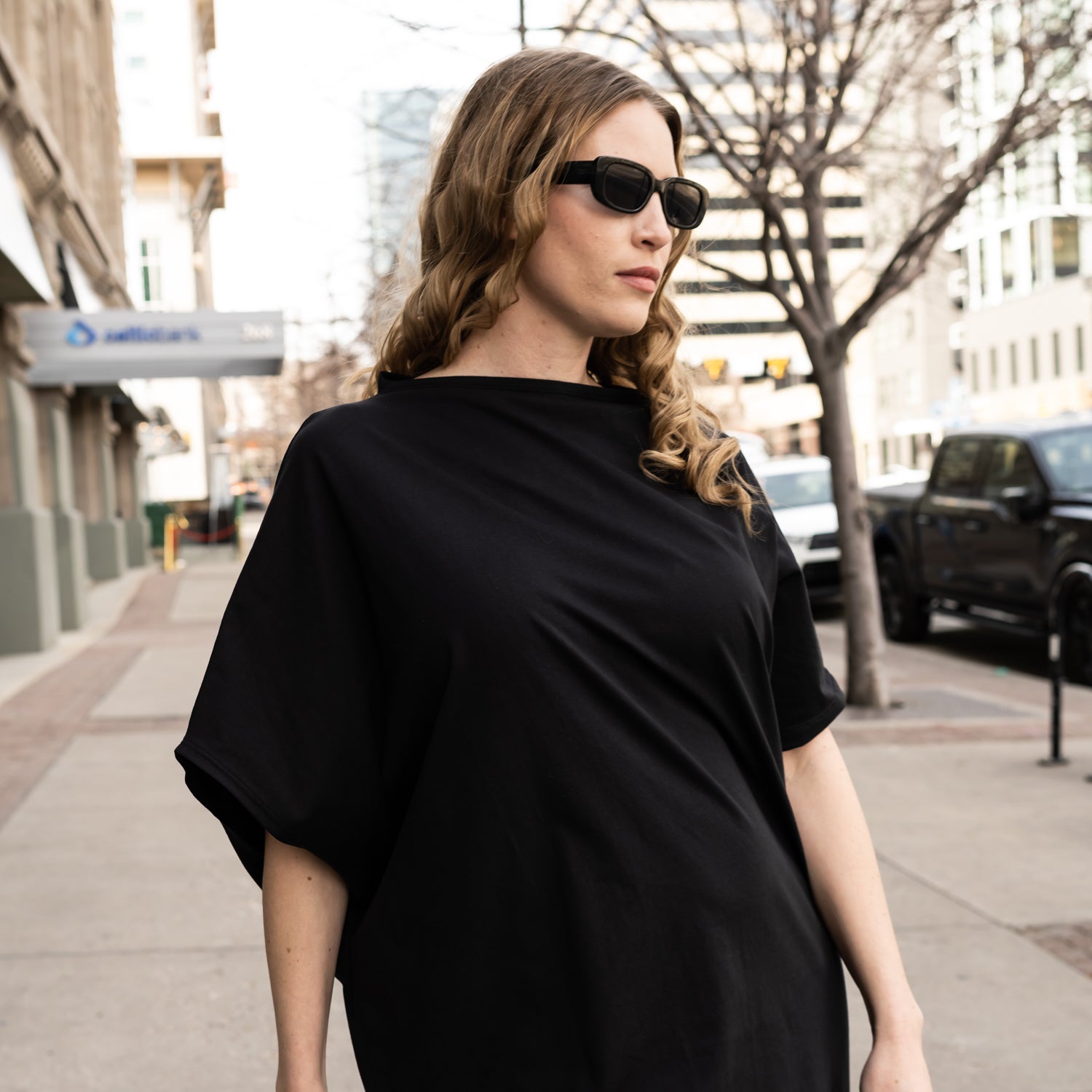 A close up of a woman wearing a black boatneck dress with asymmetrical sleeves and hem by Malaika New York