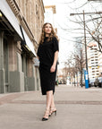 A woman wearing a long black shift dress and high heels. A long asymmetrical beautifully draped dress by Malaika New York