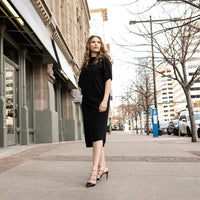 A woman wearing a long black shift dress and high heels. A long asymmetrical beautifully draped dress by Malaika New York
