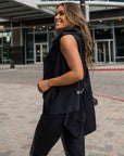 A side view of a woman smiling wearing Malaika New York. She is wearing a asymmetrical black vest with old bike tube details & vegan leather leggings