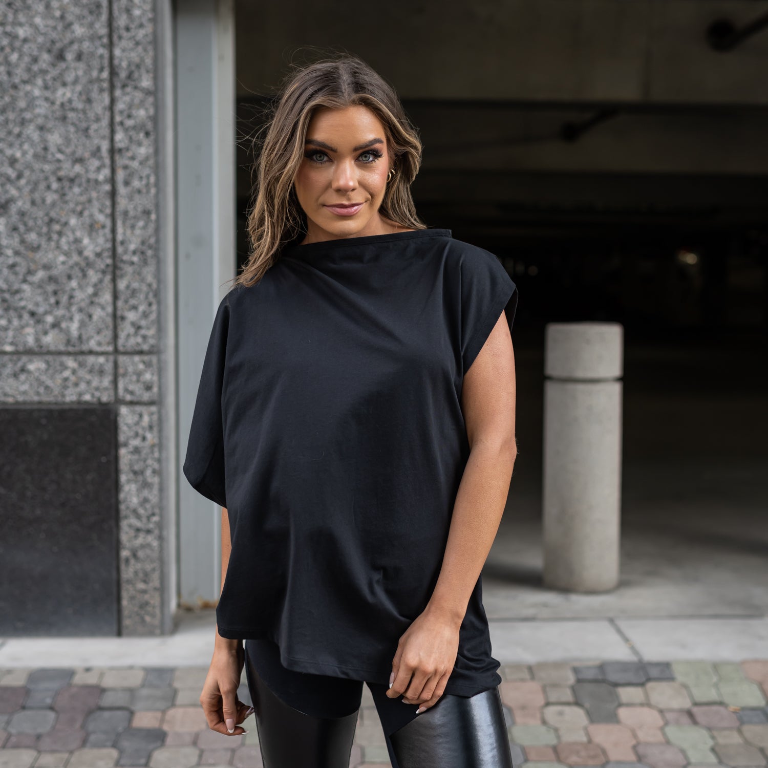 A close up of a woman wearing an asymmetrical black organic cotton t-shirt and a pair of faux leather leggings by Malaika New York