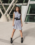 A women is wearing a grey organic cotton  knee length dress with a belt in front of a building 